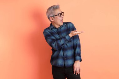 Yeşil ekose elbiseli, gözlüklü, turuncu arka planda izole edilmiş bir yere işaret eden Asyalı son sınıf öğrencisi. Turist, akıllı gündelik.