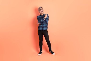 Yeşil ekose giysili Asyalı son sınıf öğrencisi gözlüklü, ayakta duran ve turuncu arka planda sırıtan. Turist, akıllı gündelik.