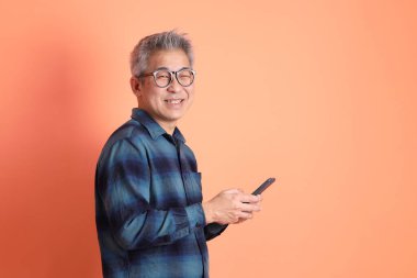 Yeşil ekose elbiseli Asyalı son sınıf öğrencisi gözlüklü turuncu arka planda izole edilmiş cep telefonu jesti. Turist, akıllı gündelik.