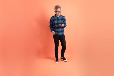 Yeşil ekose elbiseli Asyalı son sınıf öğrencisi gözlüklü turuncu arka planda izole edilmiş cep telefonu jesti. Turist, akıllı gündelik.