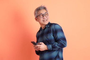 Yeşil ekose elbiseli Asyalı son sınıf öğrencisi gözlüklü turuncu arka planda izole edilmiş cep telefonu jesti. Turist, akıllı gündelik.