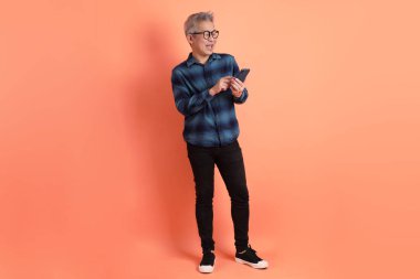 Yeşil ekose elbiseli Asyalı son sınıf öğrencisi gözlüklü turuncu arka planda izole edilmiş cep telefonu jesti. Turist, akıllı gündelik.