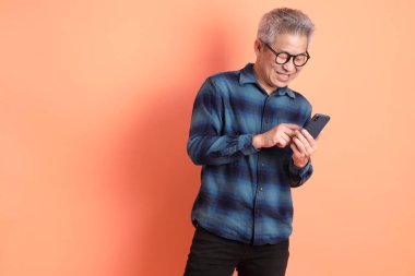 Yeşil ekose elbiseli Asyalı son sınıf öğrencisi gözlüklü turuncu arka planda izole edilmiş cep telefonu jesti. Turist, akıllı gündelik.