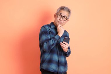 Yeşil ekose elbiseli, gözlüklü, cep telefonu tutan ve turuncu arka planda izole edilmiş olduğunu düşünen Asyalı son sınıf öğrencisi. Turist, akıllı gündelik.