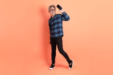 Yeşil ekose elbiseli Asyalı son sınıf öğrencisi gözlüklü turuncu arka planda izole edilmiş cep telefonu jesti. Turist, akıllı gündelik.