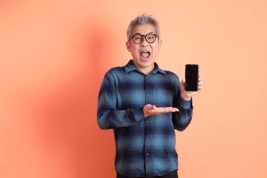 Yeşil ekose elbiseli, gözlüklü, cep telefonu tutan ve turuncu arka planda izole edilmiş bir yere işaret eden Asyalı son sınıf öğrencisi. Turist, akıllı gündelik.