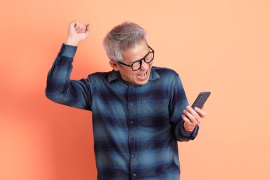 Yeşil ekose elbiseli Asyalı son sınıf öğrencisi cep telefonu jestli gözlük takıyor ve evet, heyecanlı, turuncu arka planda izole edilmiş telefonu kutluyor. Turist, akıllı gündelik.