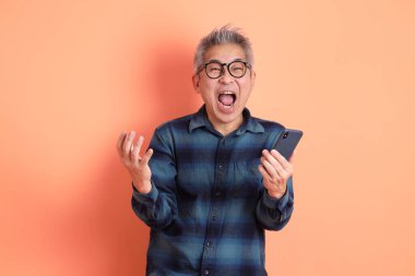 Yeşil ekose elbiseli Asyalı son sınıf öğrencisi cep telefonu ve sürpriz hareketleriyle gözlük takıyor ve turuncu arka planda etkileyici bir şekilde izole edilmiş. Turist, akıllı gündelik.