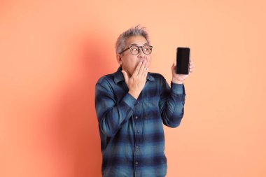 Yeşil ekose elbiseli Asyalı son sınıf öğrencisi cep telefonu ve sürpriz hareketleriyle gözlük takıyor ve turuncu arka planda etkileyici bir şekilde izole edilmiş. Turist, akıllı gündelik.