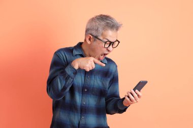 Yeşil ekose elbiseli Asyalı son sınıf öğrencisi gözlüklü turuncu arka planda izole edilmiş cep telefonu jesti. Turist, akıllı gündelik.