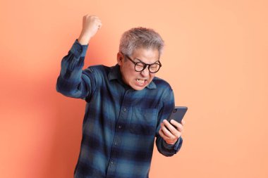 Yeşil ekose elbiseli Asyalı son sınıf öğrencisi cep telefonu jestli gözlük takıyor ve evet, heyecanlı, turuncu arka planda izole edilmiş telefonu kutluyor. Turist, akıllı gündelik.