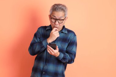 Yeşil ekose elbiseli, gözlüklü, cep telefonu tutan ve turuncu arka planda izole edilmiş olduğunu düşünen Asyalı son sınıf öğrencisi. Turist, akıllı gündelik.