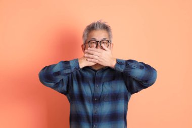 Yeşil ekose elbiseli Asyalı son sınıf öğrencisi gözlüklü, ağzı kapalı, turuncu arka planda izole edilmiş sessiz bir jest. Turist, akıllı gündelik.