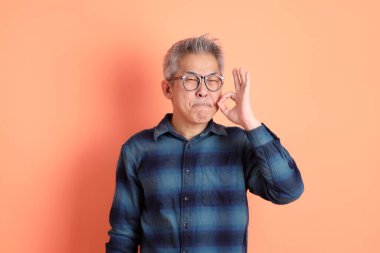 Yeşil ekose elbiseli Asyalı son sınıf öğrencisi gözlüklü, ağzı kapalı, turuncu arka planda izole edilmiş sessiz bir jest. Turist, akıllı gündelik.