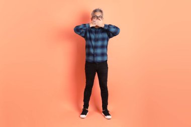 Yeşil ekose elbiseli Asyalı son sınıf öğrencisi gözlüklü, ağzı kapalı, turuncu arka planda izole edilmiş sessiz bir jest. Turist, akıllı gündelik.