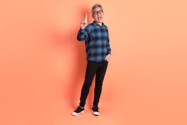 Yeşil ekose elbiseli, gözlüklü, turuncu arka planda izole edilmiş bir yere işaret eden Asyalı son sınıf öğrencisi. Turist, akıllı gündelik.