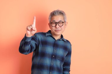 Yeşil ekose elbiseli, gözlüklü, turuncu arka planda izole edilmiş bir yere işaret eden Asyalı son sınıf öğrencisi. Turist, akıllı gündelik.