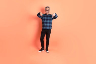 Yeşil ekose elbiseli, gözlüklü, turuncu arka planda izole edilmiş bir yere işaret eden Asyalı son sınıf öğrencisi. Turist, akıllı gündelik.