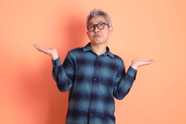 Yeşil ekose elbiseli, gözlüklü, turuncu arka planda izole edilmiş bir yere işaret eden Asyalı son sınıf öğrencisi. Turist, akıllı gündelik.