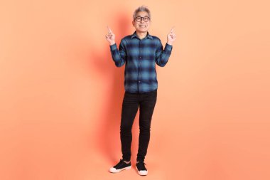 Yeşil ekose elbiseli, gözlüklü, turuncu arka planda izole edilmiş bir yere işaret eden Asyalı son sınıf öğrencisi. Turist, akıllı gündelik.