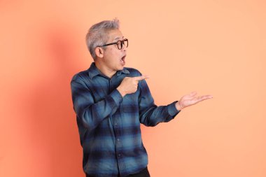 Yeşil ekose elbiseli, gözlüklü, turuncu arka planda izole edilmiş bir yere işaret eden Asyalı son sınıf öğrencisi. Turist, akıllı gündelik.