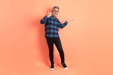 Yeşil ekose elbiseli, gözlüklü, turuncu arka planda izole edilmiş bir yere işaret eden Asyalı son sınıf öğrencisi. Turist, akıllı gündelik.