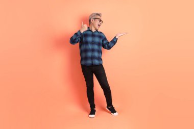 Yeşil ekose elbiseli, gözlüklü, turuncu arka planda izole edilmiş bir yere işaret eden Asyalı son sınıf öğrencisi. Turist, akıllı gündelik.