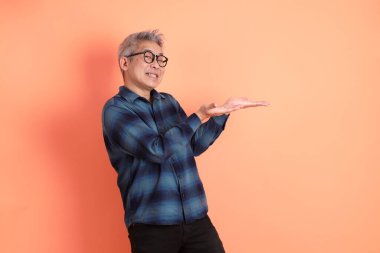 Yeşil ekose elbiseli, gözlüklü, turuncu arka planda izole edilmiş bir yere işaret eden Asyalı son sınıf öğrencisi. Turist, akıllı gündelik.