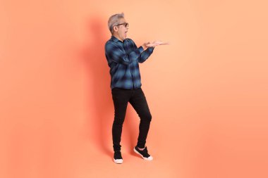 Yeşil ekose elbiseli, gözlüklü, turuncu arka planda izole edilmiş bir yere işaret eden Asyalı son sınıf öğrencisi. Turist, akıllı gündelik.