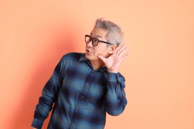 Yeşil ekose elbiseli Asyalı son sınıf öğrencisi gözlük takıyor ve turuncu arka planda izole edilmiş dinleme ya da işitme hareketi yapıyor. Turist, akıllı gündelik.