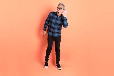 Yeşil ekose elbiseli Asyalı son sınıf öğrencisi gözlük takıyor ve turuncu arka planda izole edilmiş dinleme ya da işitme hareketi yapıyor. Turist, akıllı gündelik.