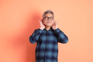 Yeşil ekose elbiseli Asyalı son sınıf öğrencisi gözlük takıyor ve turuncu arka planda izole edilmiş dinleme ya da işitme hareketi yapıyor. Turist, akıllı gündelik.