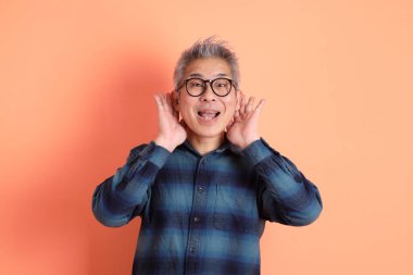 Yeşil ekose elbiseli Asyalı son sınıf öğrencisi gözlük takıyor ve turuncu arka planda izole edilmiş dinleme ya da işitme hareketi yapıyor. Turist, akıllı gündelik.