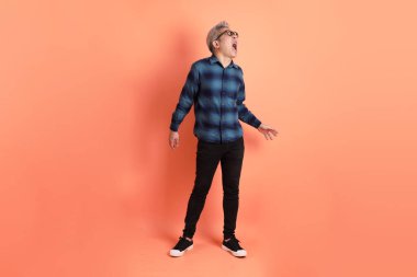 Yeşil ekose giysili Asyalı son sınıf öğrencisi. Gözlük takıyor. Bağırıyor, elinde turuncu arka planda megafon tutuyor. Turist, akıllı gündelik.