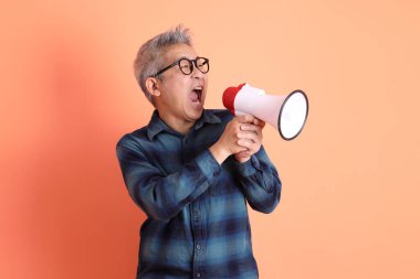 Yeşil ekose giysili Asyalı son sınıf öğrencisi. Gözlük takıyor. Bağırıyor, elinde turuncu arka planda megafon tutuyor. Turist, akıllı gündelik.