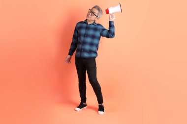 Yeşil ekose giysili Asyalı son sınıf öğrencisi. Gözlük takıyor. Bağırıyor, elinde turuncu arka planda megafon tutuyor. Turist, akıllı gündelik.