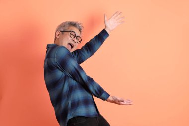 Yeşil ekose elbiseli, gözlüklü, turuncu arka planda izole edilmiş bir yere işaret eden Asyalı son sınıf öğrencisi. Turist, akıllı gündelik.