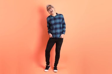 Yeşil ekose giysili Asyalı son sınıf öğrencisi gözlüklü, ayakta duran ve turuncu arka planda sırıtan. Turist, akıllı gündelik.