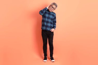 Yeşil ekose giysili Asyalı son sınıf öğrencisi, turuncu arka planda izole edilmiş kulak karıştırma hareketi yapan gözlük takıyor. Turist, akıllı gündelik.