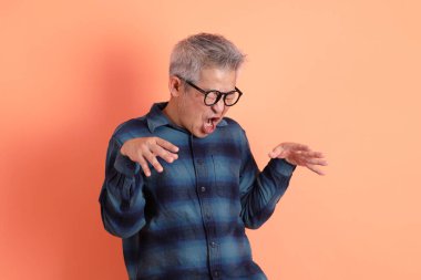 Yeşil ekose giysili Asyalı son sınıf öğrencisi gözlük takıyor ve turuncu arka planda hastalıklı davranışlar sergiliyor. Turist, akıllı gündelik.