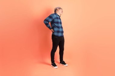 Yeşil ekose giysili Asyalı son sınıf öğrencisi. Gözlük takıyor. Sırt ağrısı jesti turuncu arka planda. Turist, akıllı gündelik.