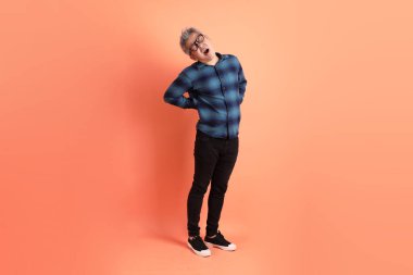 Yeşil ekose giysili Asyalı son sınıf öğrencisi. Gözlük takıyor. Sırt ağrısı jesti turuncu arka planda. Turist, akıllı gündelik.