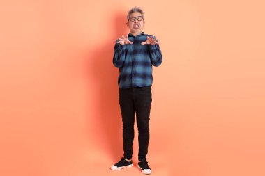 Yeşil ekose giysili Asyalı son sınıf öğrencisi gözlük takıyor ve turuncu arka planda hastalıklı davranışlar sergiliyor. Turist, akıllı gündelik.