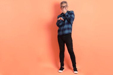 Yeşil ekose giysili Asyalı son sınıf öğrencisi gözlük takıyor ve turuncu arka planda hastalıklı davranışlar sergiliyor. Turist, akıllı gündelik.