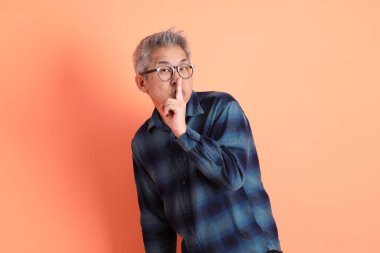 Yeşil ekose elbiseli Asyalı son sınıf öğrencisi gözlüklü, ağzı kapalı, turuncu arka planda izole edilmiş sessiz bir jest. Turist, akıllı gündelik.