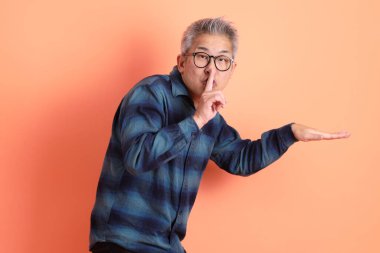 Yeşil ekose elbiseli Asyalı son sınıf öğrencisi gözlüklü, ağzı kapalı, turuncu arka planda izole edilmiş sessiz bir jest. Turist, akıllı gündelik.