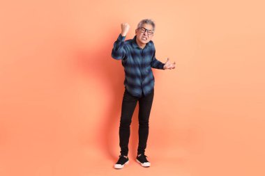 Yeşil ekose elbiseli Asyalı son sınıf öğrencisi gözlük takıyor ve turuncu arka planda öfkeli ve çığlıklar atıyor. Turist, akıllı gündelik.