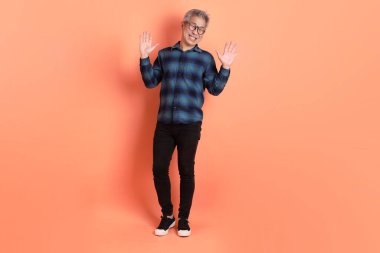 Yeşil ekose elbiseli Asyalı son sınıf öğrencisi gözlüklü turuncu arka planda izole edilmiş bir selamlama jesti takıyor. Turist, akıllı gündelik.