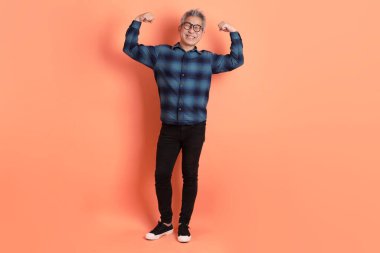 Yeşil ekose elbiseli Asyalı son sınıf öğrencisi. Gözlük takıyor. Turuncu arka planda, sağlıklı ve güçlü izole edilmiş bir jest yapıyor. Turist, akıllı gündelik.