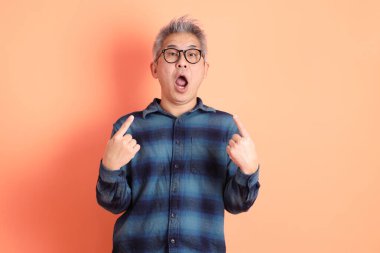 Yeşil ekose elbiseli, gözlüklü, turuncu arka planda izole edilmiş bir yere işaret eden Asyalı son sınıf öğrencisi. Turist, akıllı gündelik.